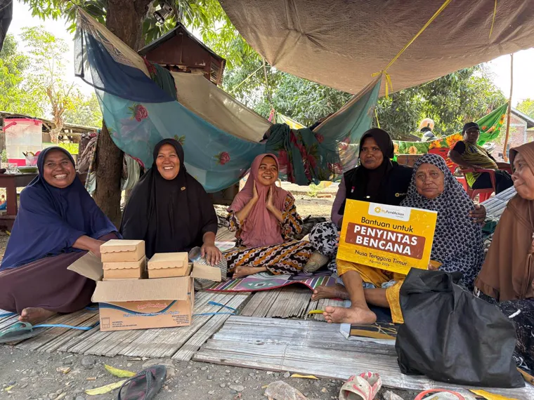 Five women sheltering under a tarpaulin in Nagekeo with a MyFundAction Bantuan untuk Penyintas Bencana card and boxes of supplies