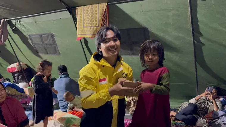A MyFundAction team member in a yellow jacket handing a hot meal to a young girl inside a shelter tent in Nagekeo