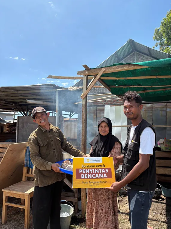 A MyFundAction team member and two residents holding a Bantuan untuk Penyintas Bencana card beside the community kitchen in Nagekeo