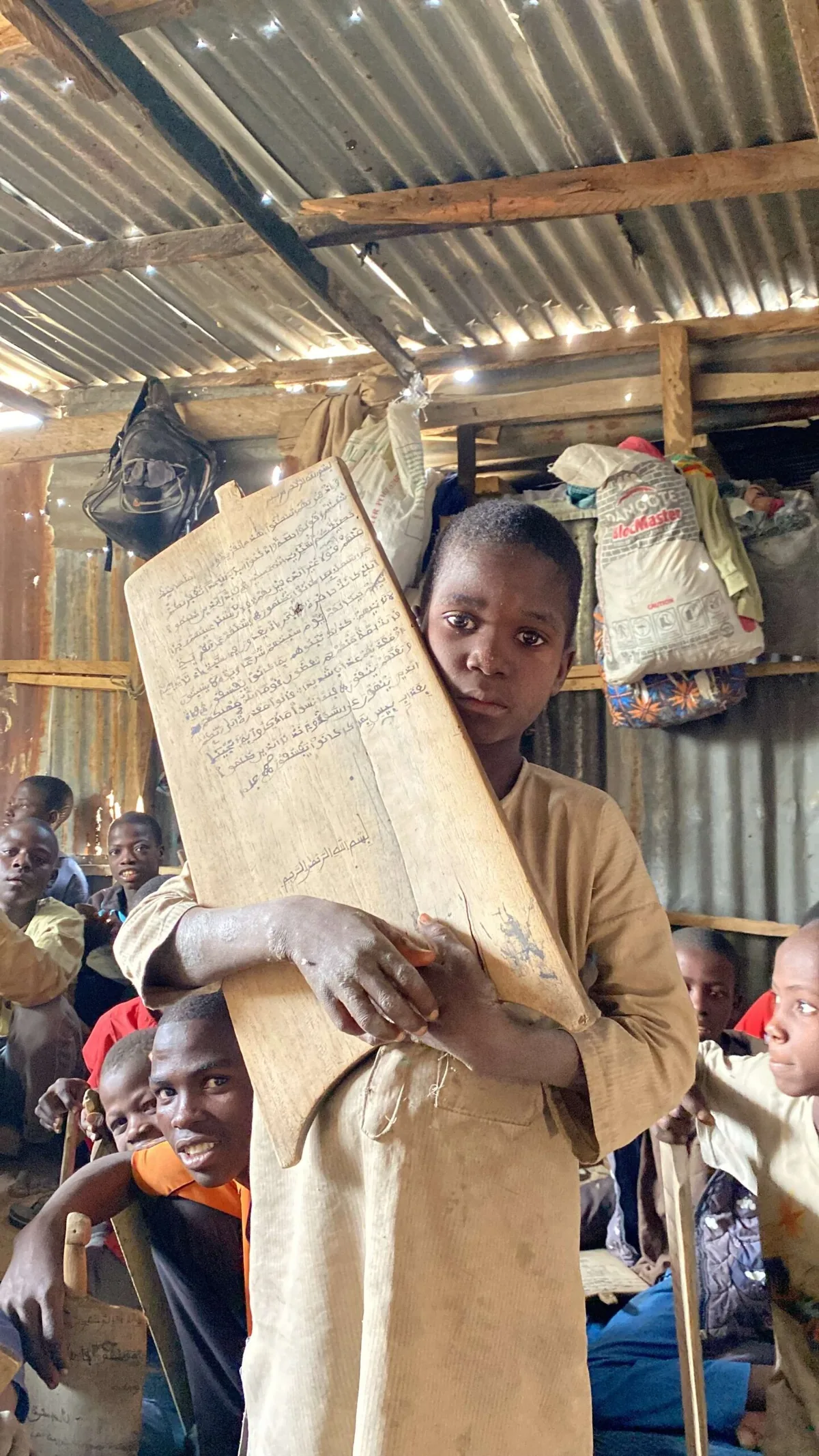 A boy in a makeshift, metal-roofed classroom in Africa holds a hand-written wooden Qur'an memorisation tablet, surrounded by other students
